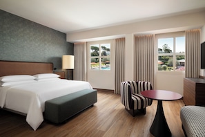 A hotel room with a bed, a desk, a chair, and a view of a pool and garden.