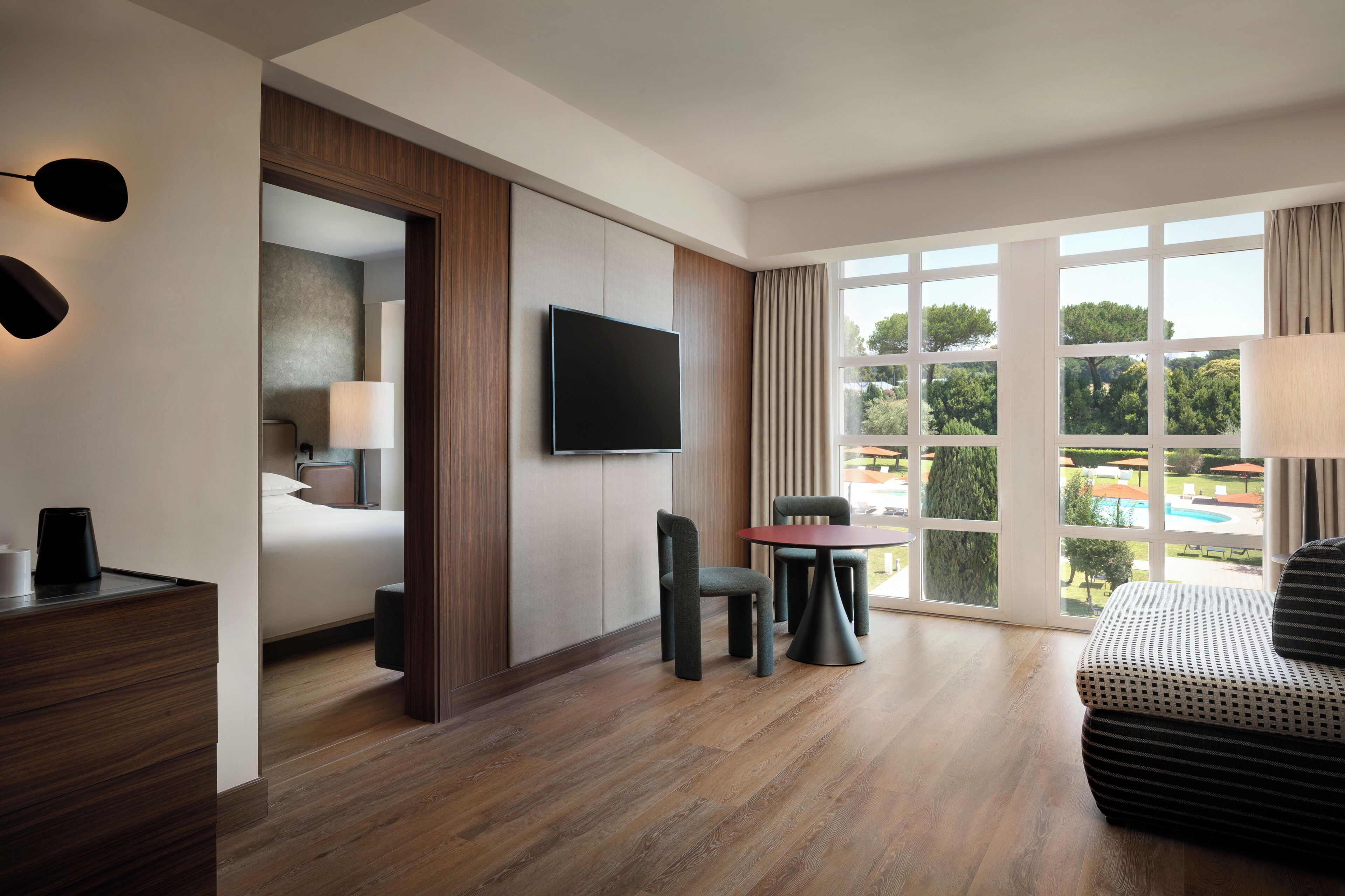 Habitación de hotel con cama, televisor montado en la pared, escritorio con lámpara y vista a una piscina a través de amplias ventanas.