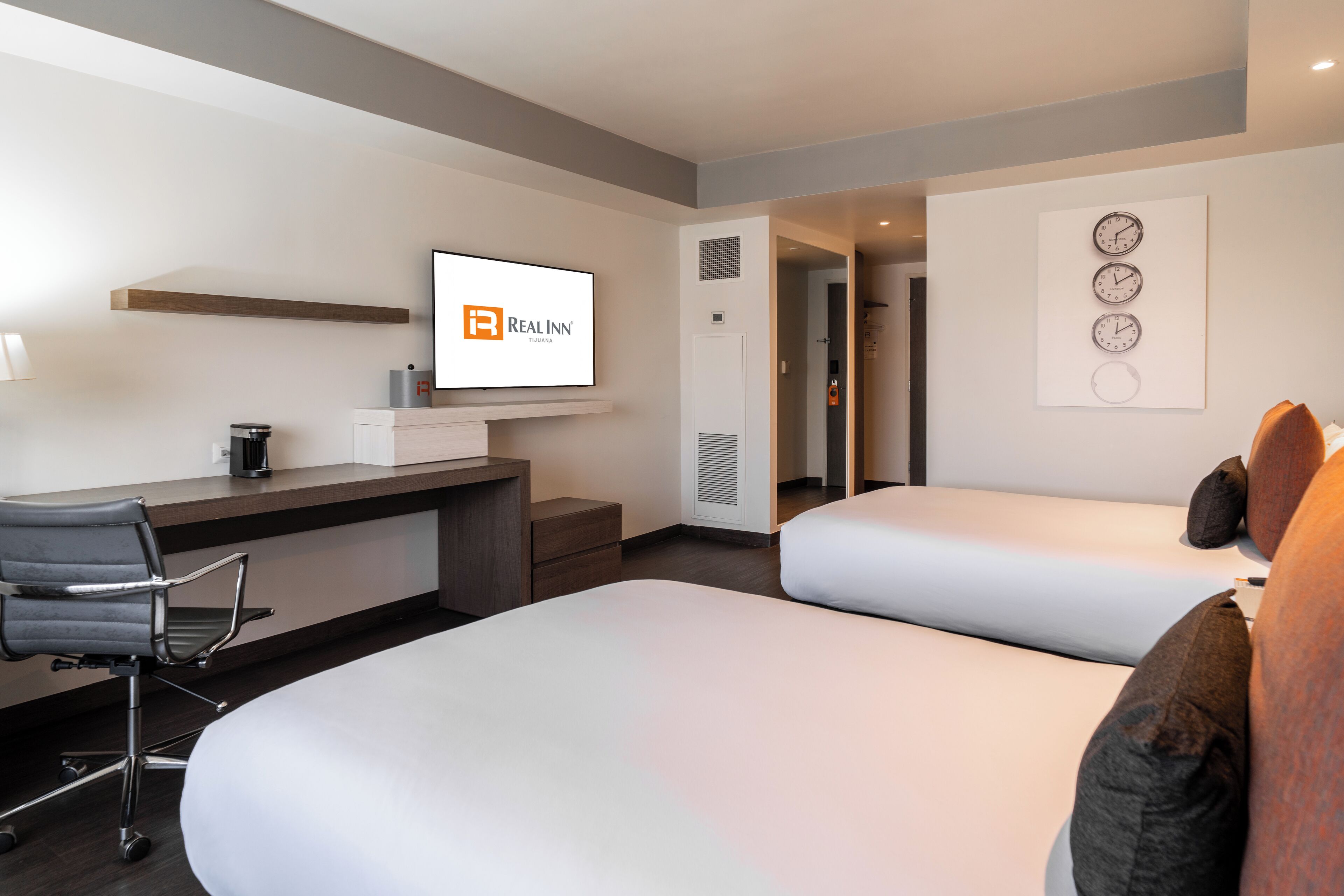 Habitación de hotel con escritorio, dos camas, televisor y un reloj en la pared.