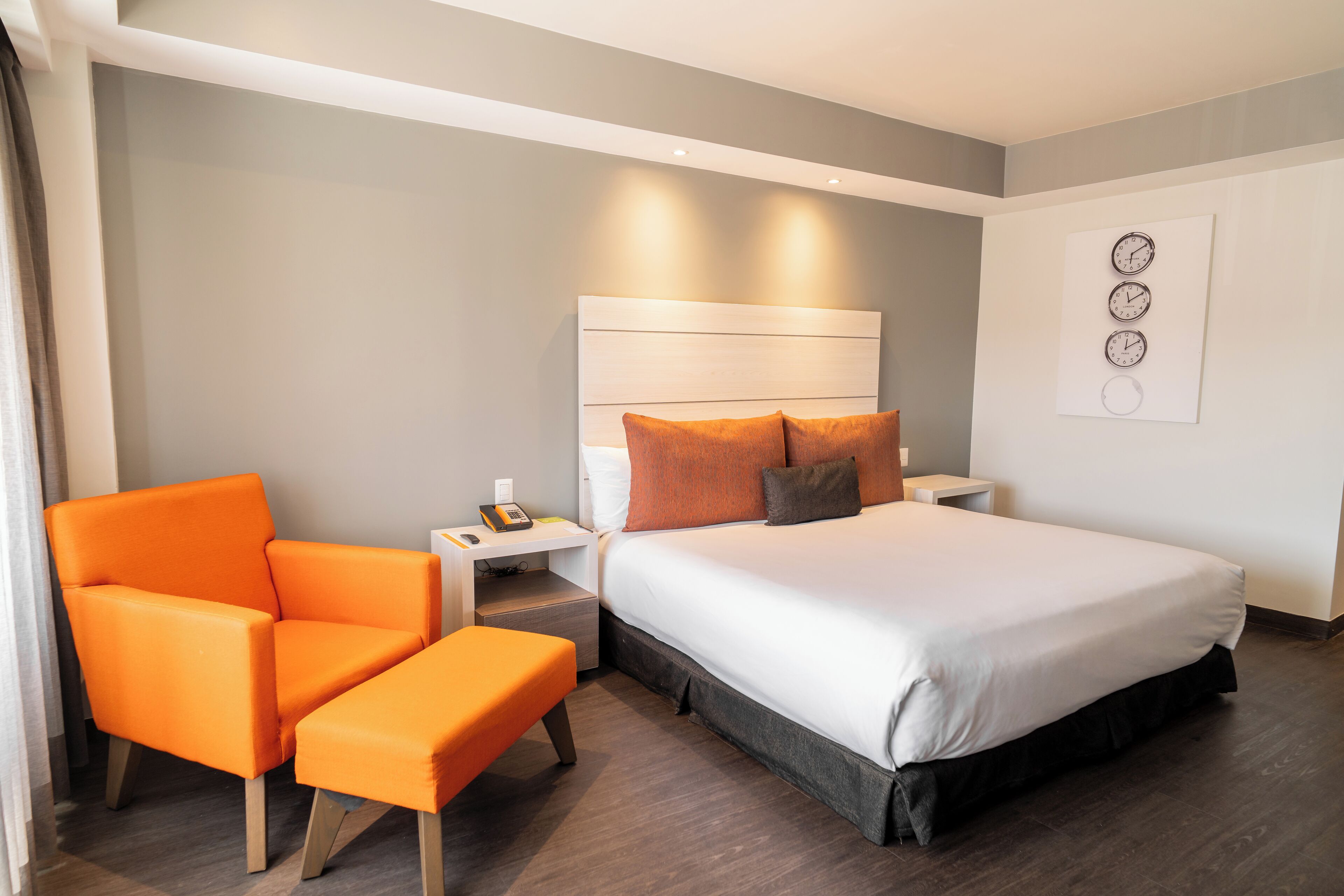 Quarto de hotel com cama, uma poltrona laranja, mesa de cabeceira e relógio de parede.