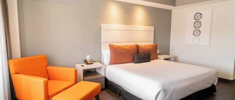 Quarto de hotel com cama, uma poltrona laranja, mesa de cabeceira e relógio de parede.