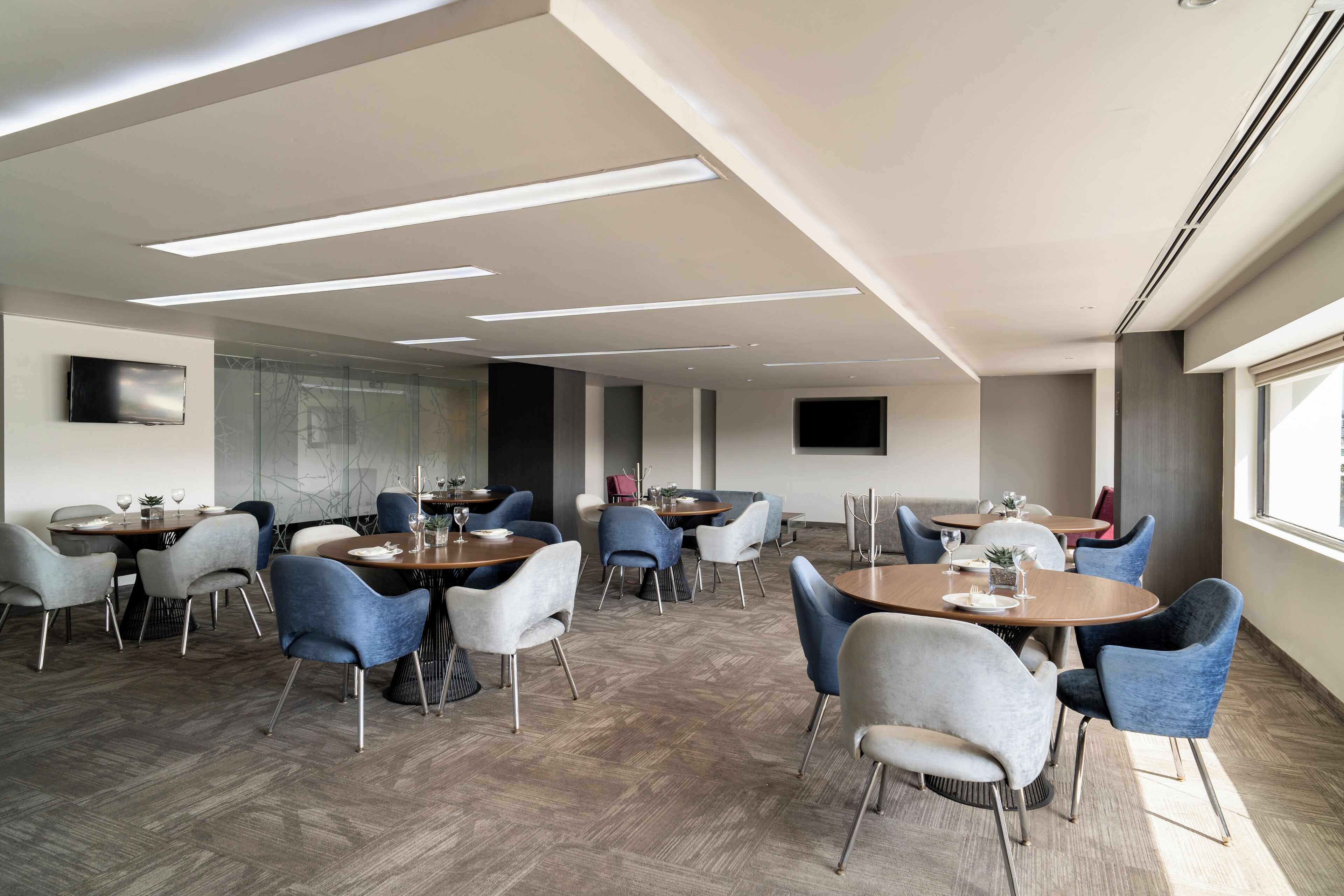 A modern dining area with multiple round tables and chairs, a herringbone floor pattern, recessed ceiling lighting, and a flat-screen TV mounted on the wall.
