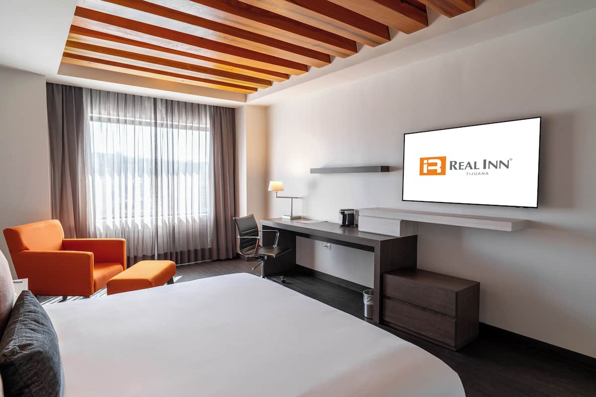 A modern hotel room with a large bed, an orange armchair, a desk with a chair, and a TV mounted on the wall displaying the Real Inn logo.