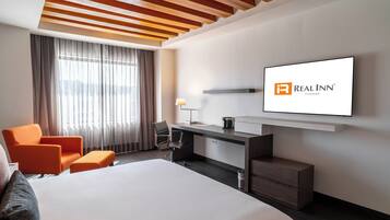 Habitación de hotel con cama, escritorio con silla, televisor montado en la pared y ventana con cortinas.