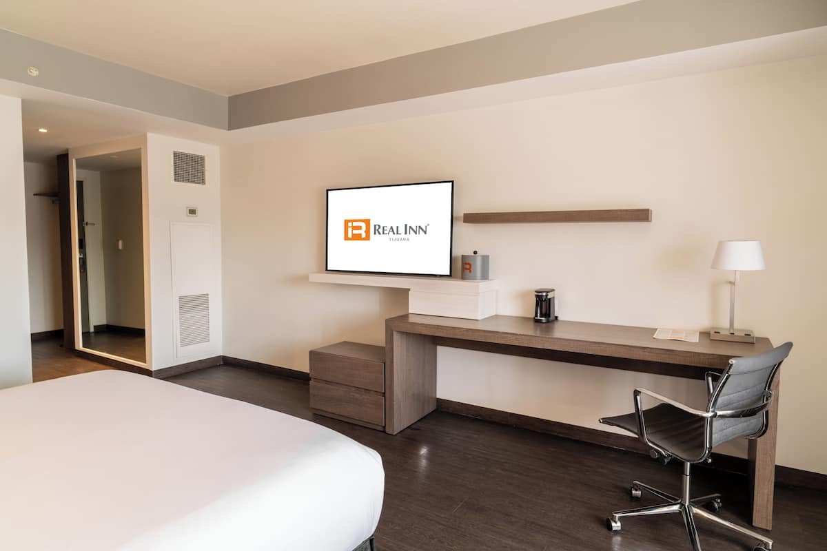 A hotel room with a bed, a desk with a chair, a TV mounted on the wall, and a door leading to another room.