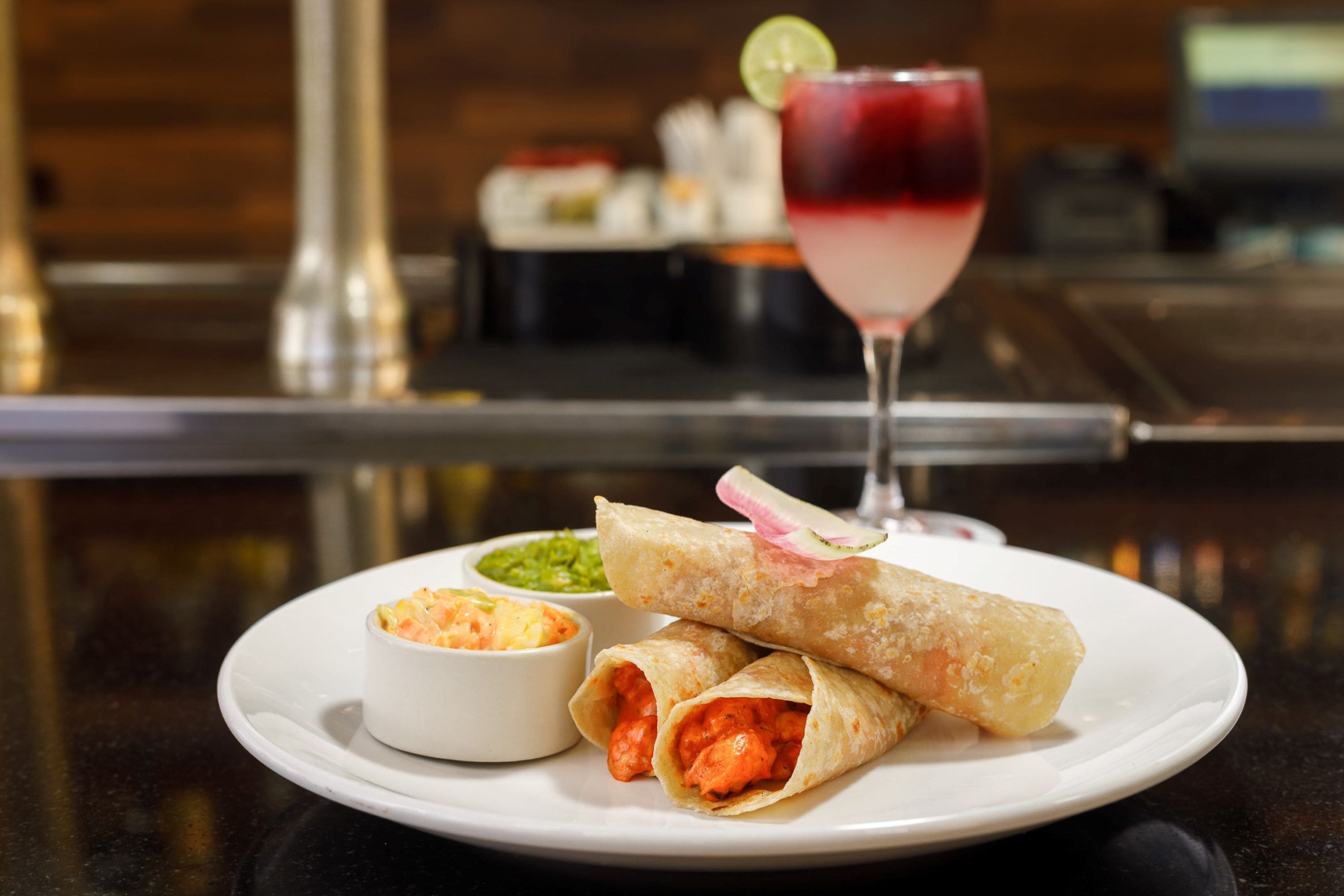 A plate with two wraps, a small bowl of sauce, and a glass of red drink on a table.