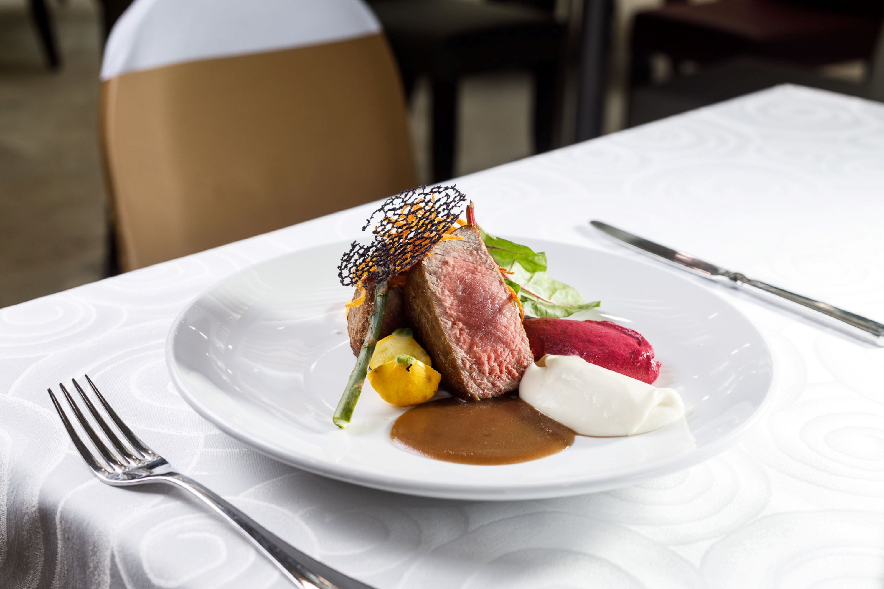 A white plate with a piece of meat, vegetables, and a sauce on a tablecloth.