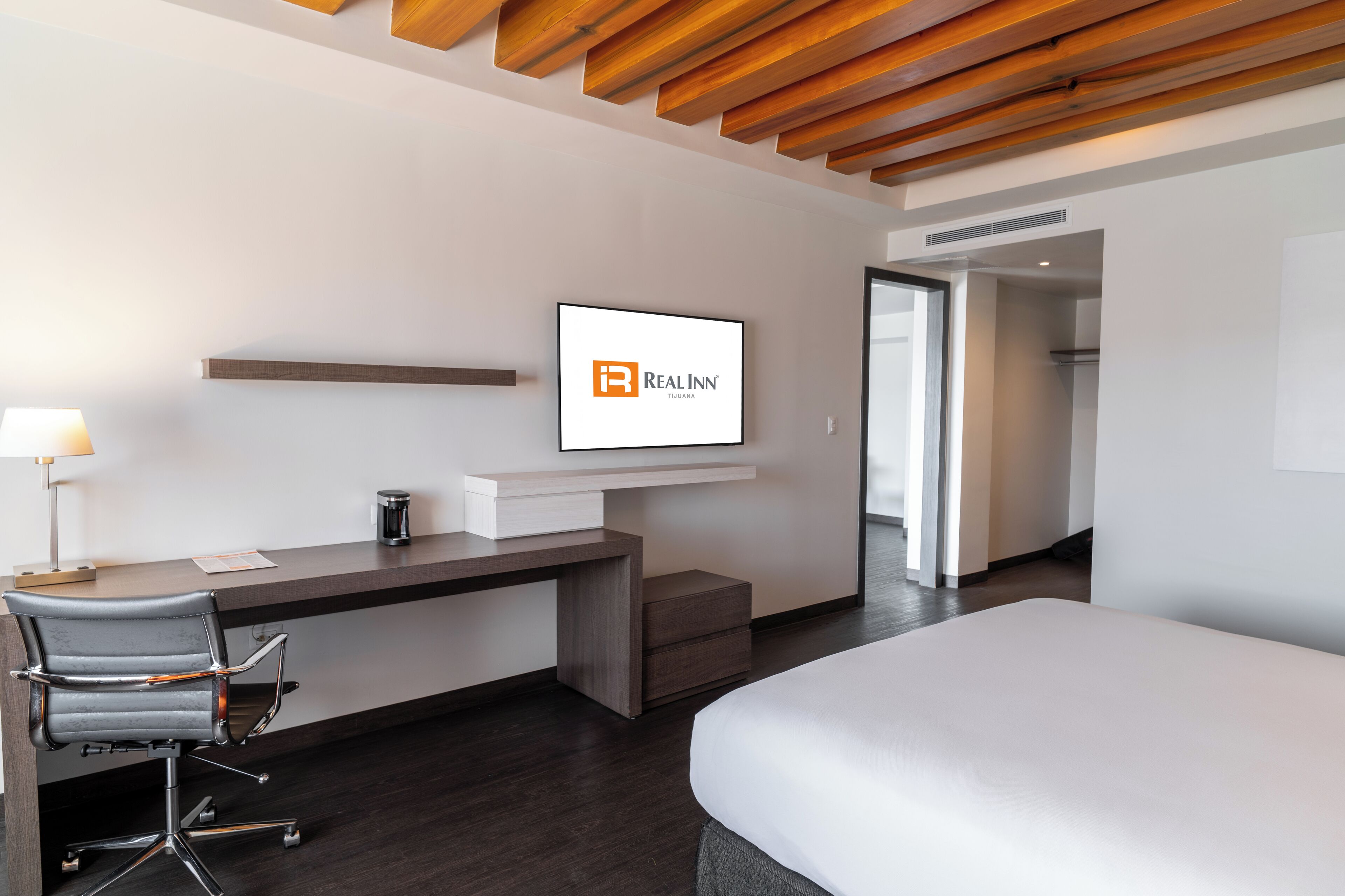 Hotelkamer met een bed, een bureau met stoel, een aan de muur bevestigde televisie en een houten plafond.