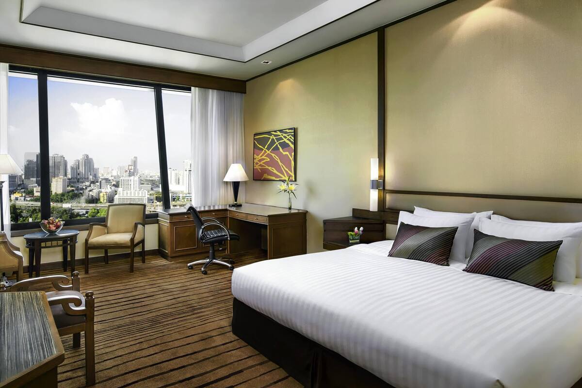 Une chambre d’hôtel avec un grand lit, un bureau, une chaise et une vue sur le paysage urbain grâce à de grandes fenêtres.