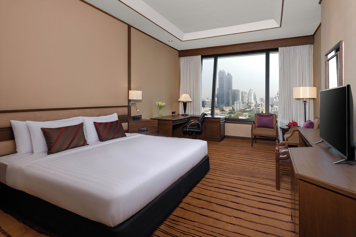 A hotel room with a large bed, a desk, a chair, a TV, and a view of the cityscape.