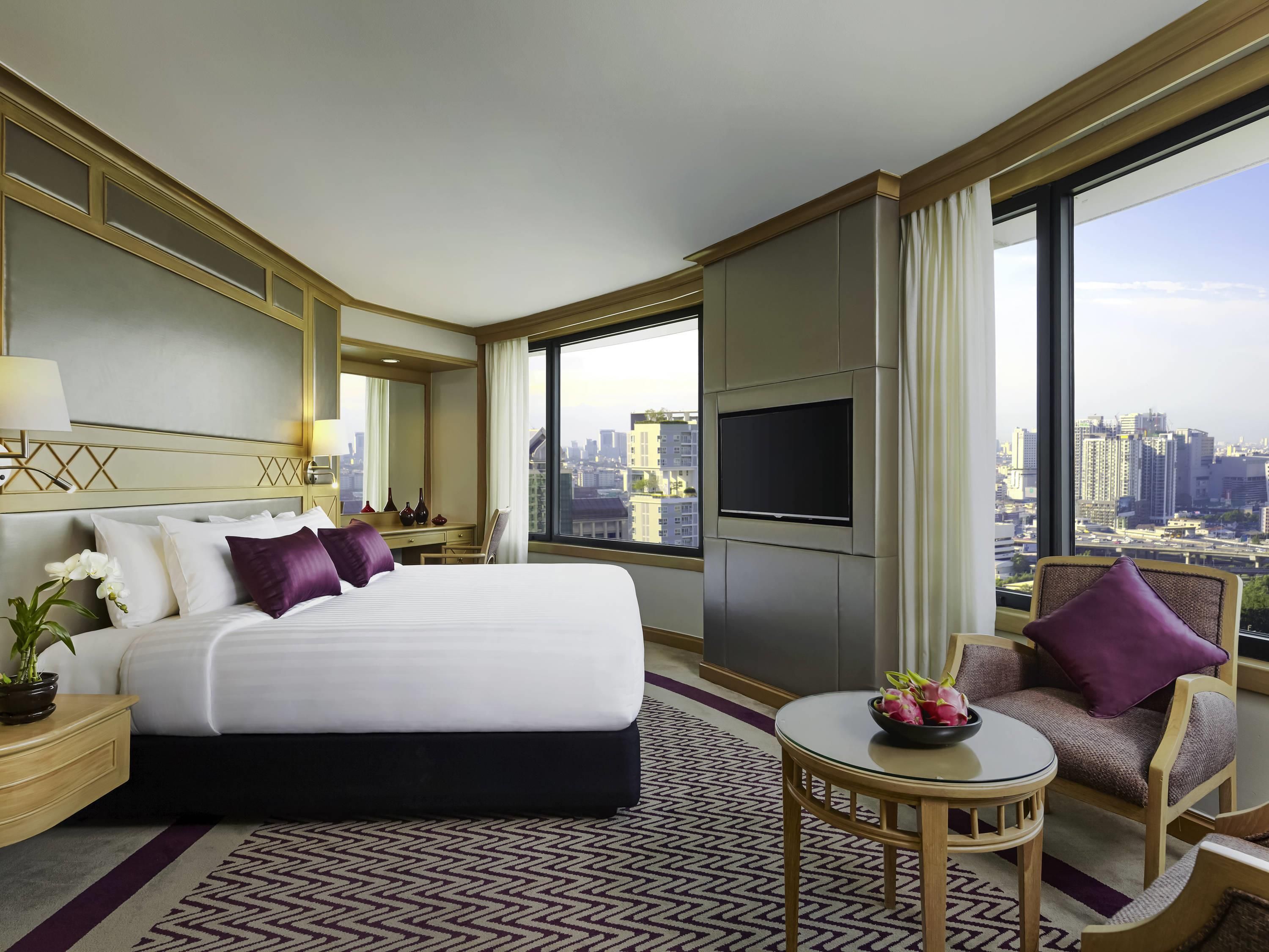 A hotel room with a large bed, a TV, a city view, and a small table with a bowl of fruit.