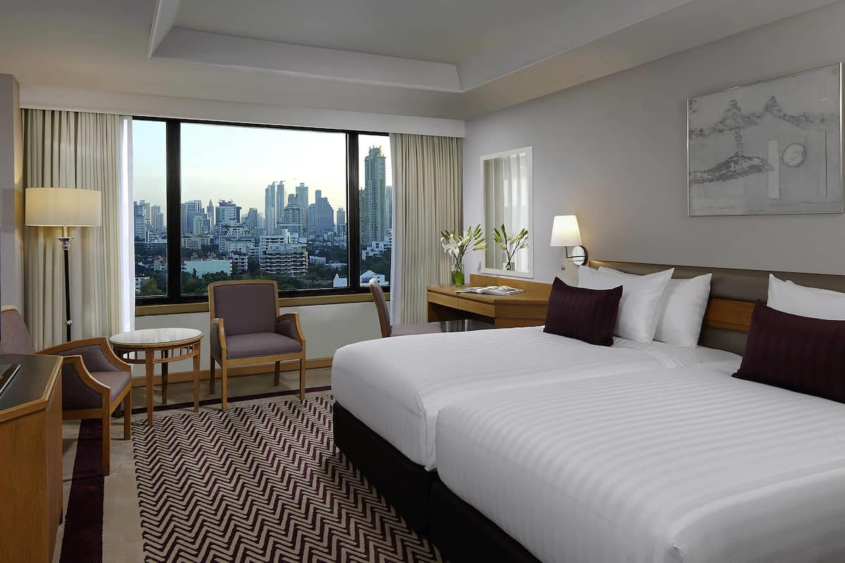 Une chambre d’hôtel avec un grand lit, un bureau, une chaise et une vue sur le paysage urbain, visible à travers une grande fenêtre.