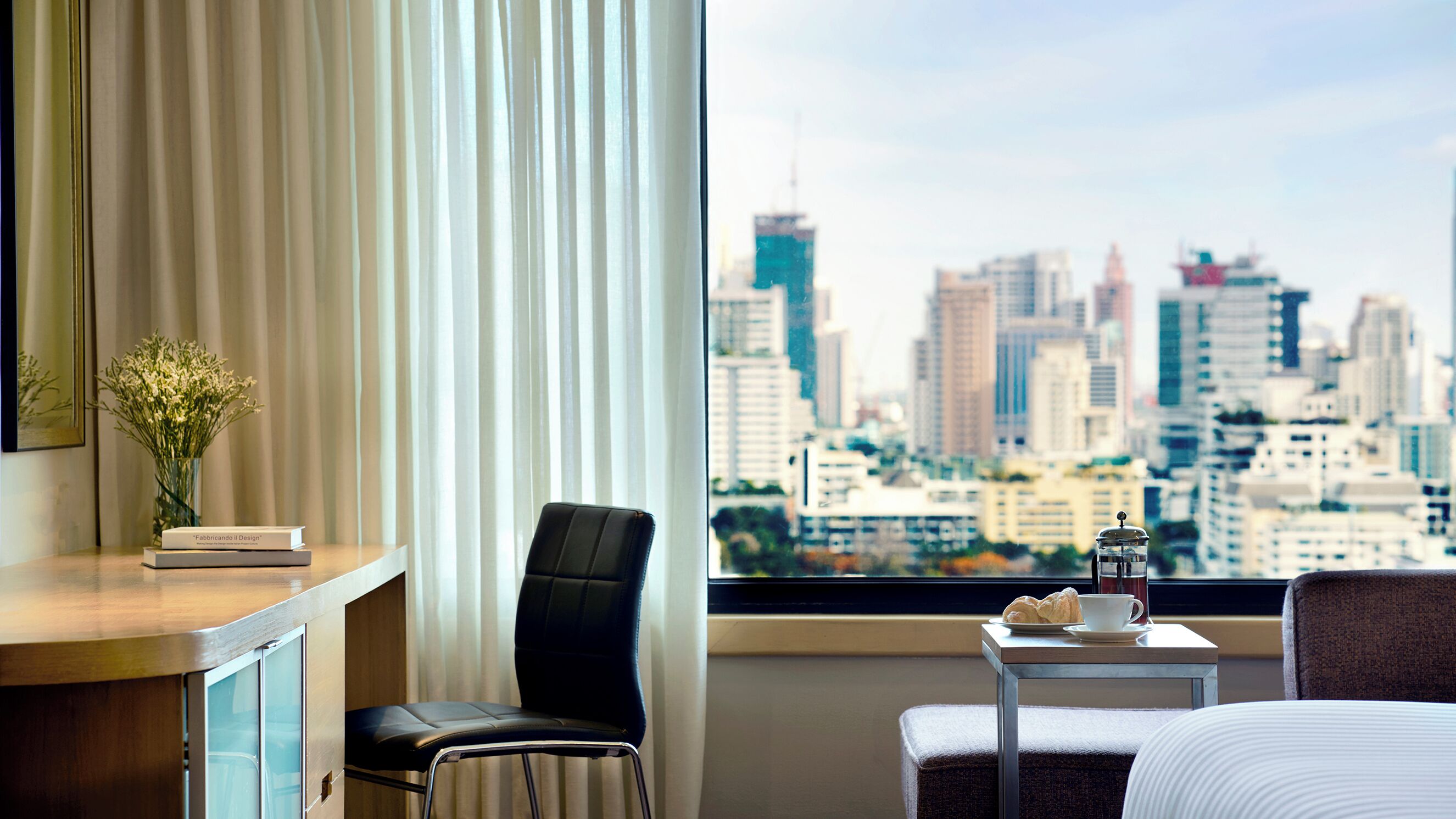 A hotel room with a city view, a desk with a vase of flowers, a chair, a small table with a cup and pastries, and a bed.