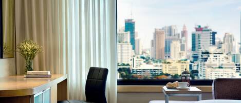 A hotel room with a city view, a desk with a vase of flowers, a chair, a small table with a cup and pastries, and a bed.