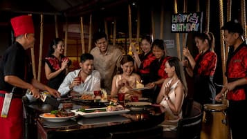 A group of people celebrating around a table with food and drinks, a neon sign reading 'HAPPY BIRTHDAY,' and a 'NO SMOKING' sign.