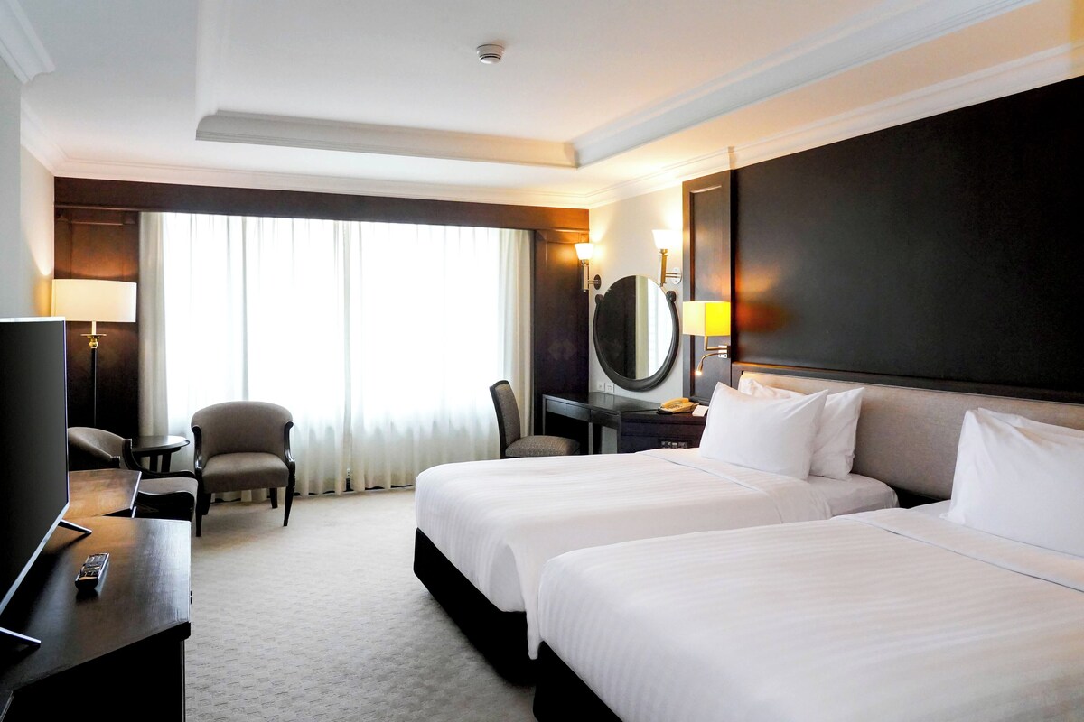 A hotel room with two beds, a desk with a TV, a chair, a desk lamp, and a large mirror.