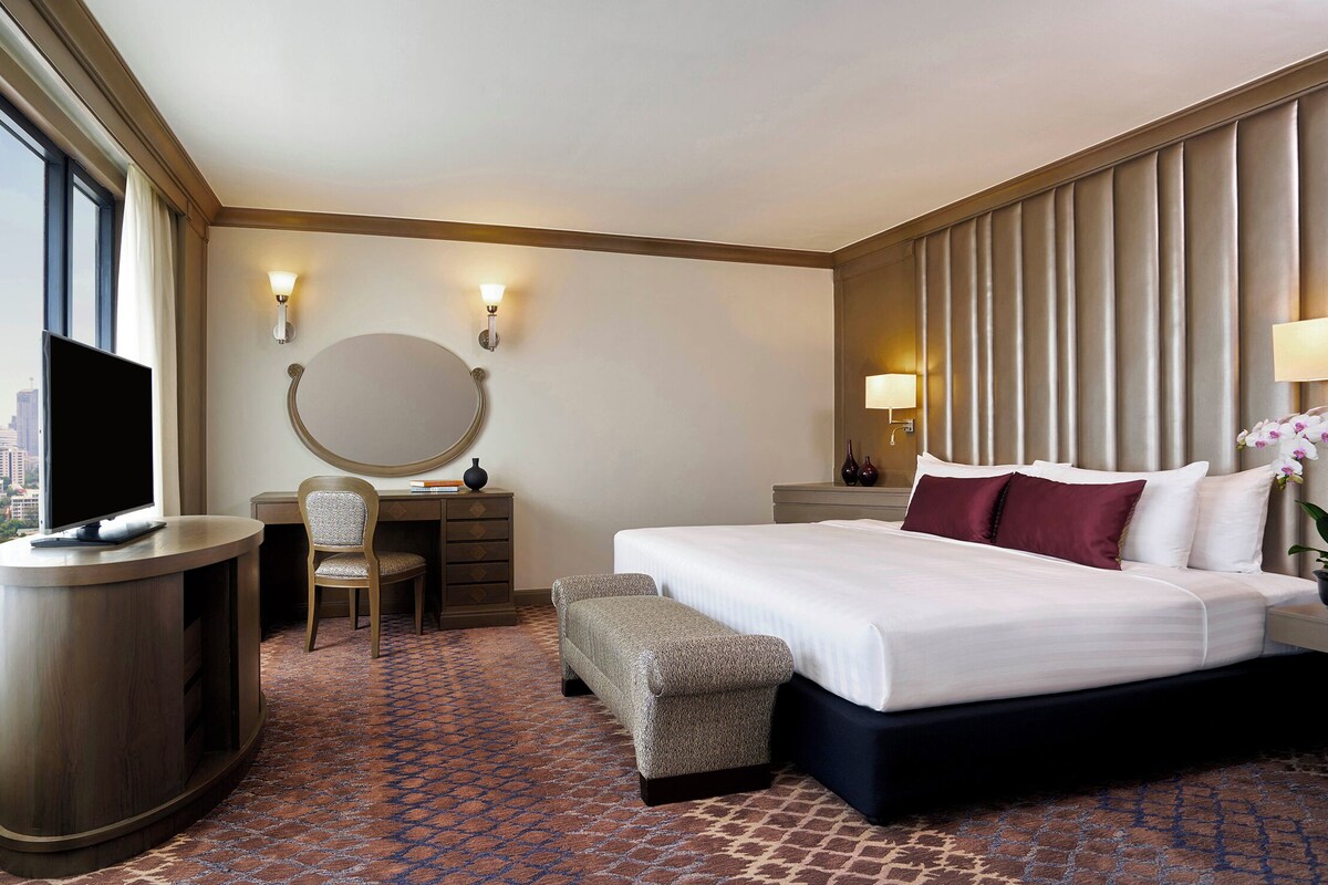 A hotel room with a large bed, a desk with a chair, a round mirror, a small bench, and a view of the city through the window.