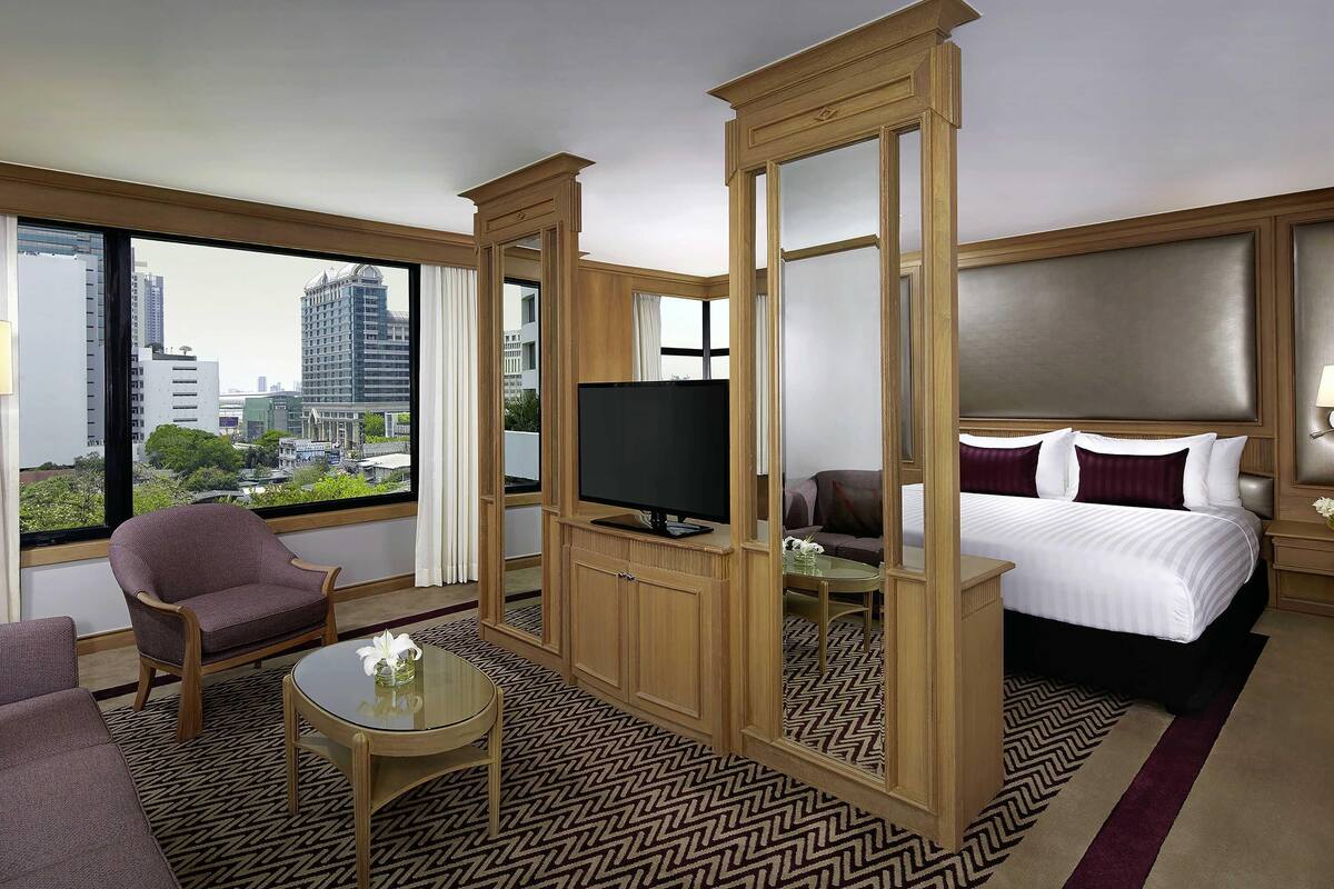 Une chambre d’hôtel équipée d’un lit, d’une télévision, d’un coin salon et d’une grande fenêtre donnant sur la ville.