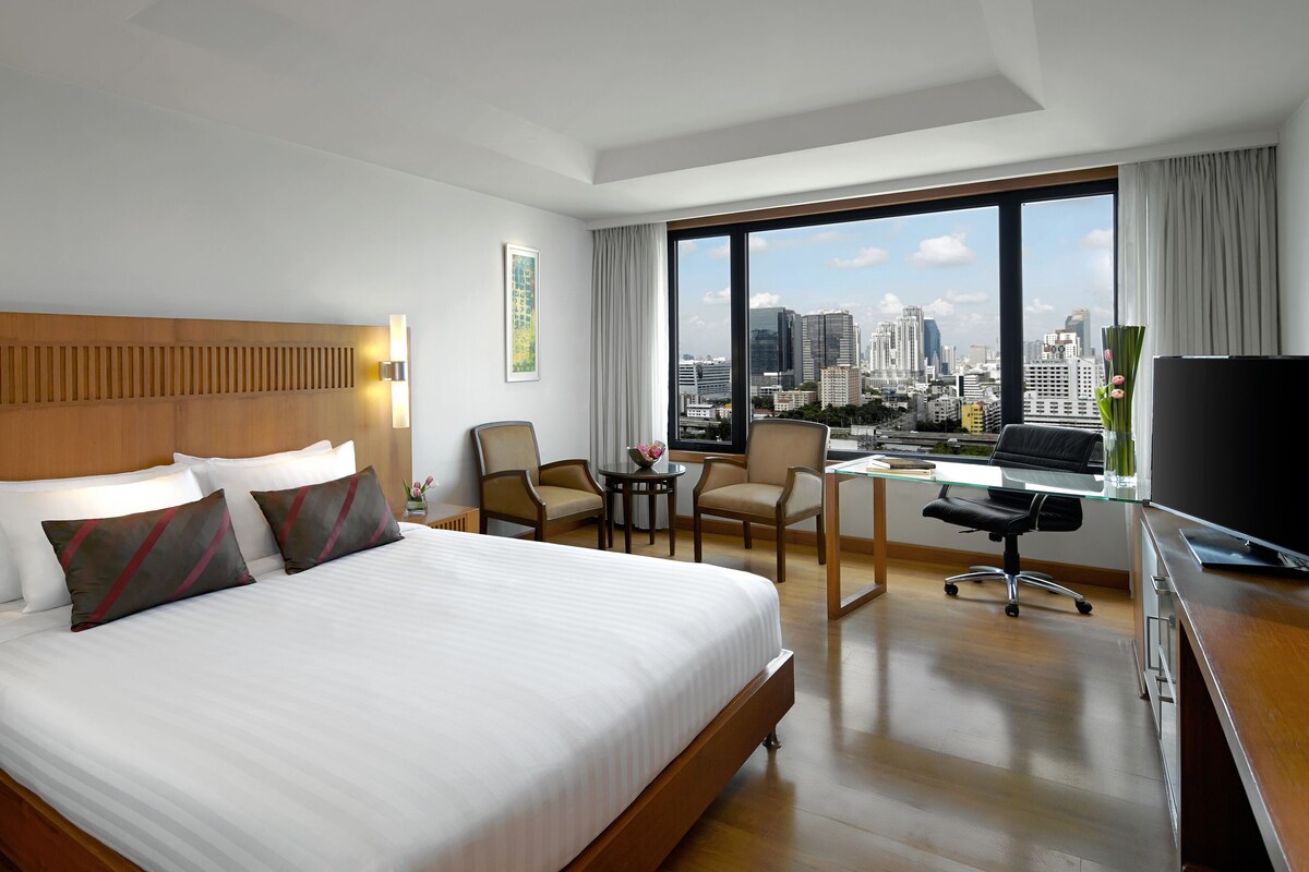 A hotel room with a large bed, a desk with a TV, a chair, and a view of the cityscape.