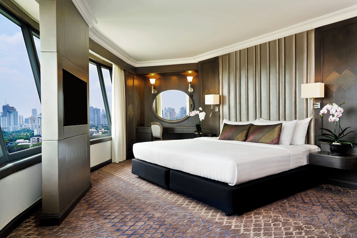 A hotel room with a large bed, a city view, a round mirror, and two wall-mounted lamps.