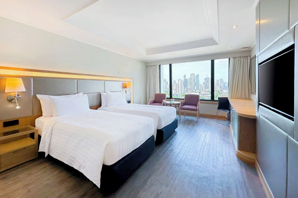 A hotel room with two beds, a desk, chairs, a TV, and a city view.