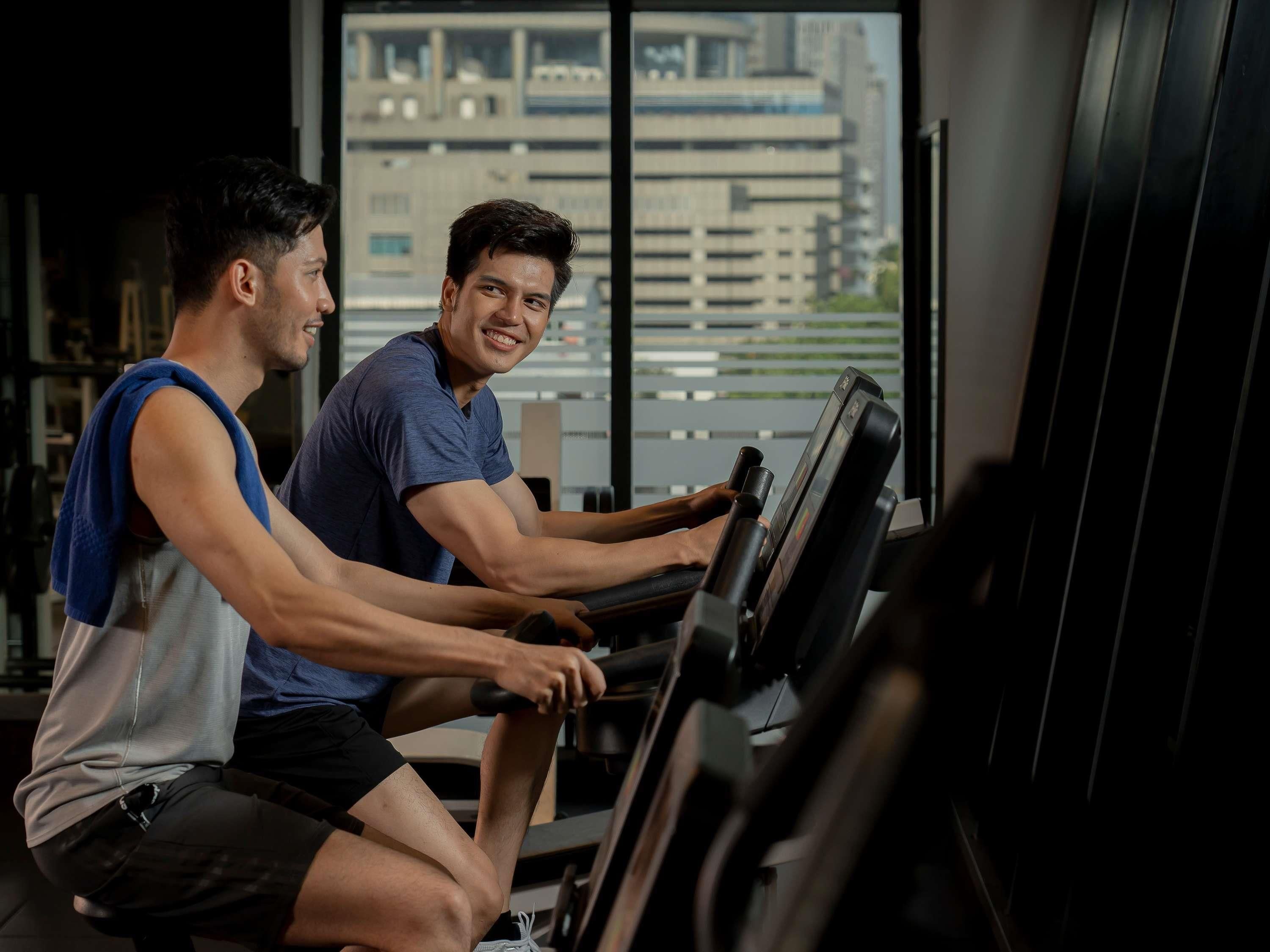 Two individuals using a stationary bike in a gym with a cityscape view through large windows.