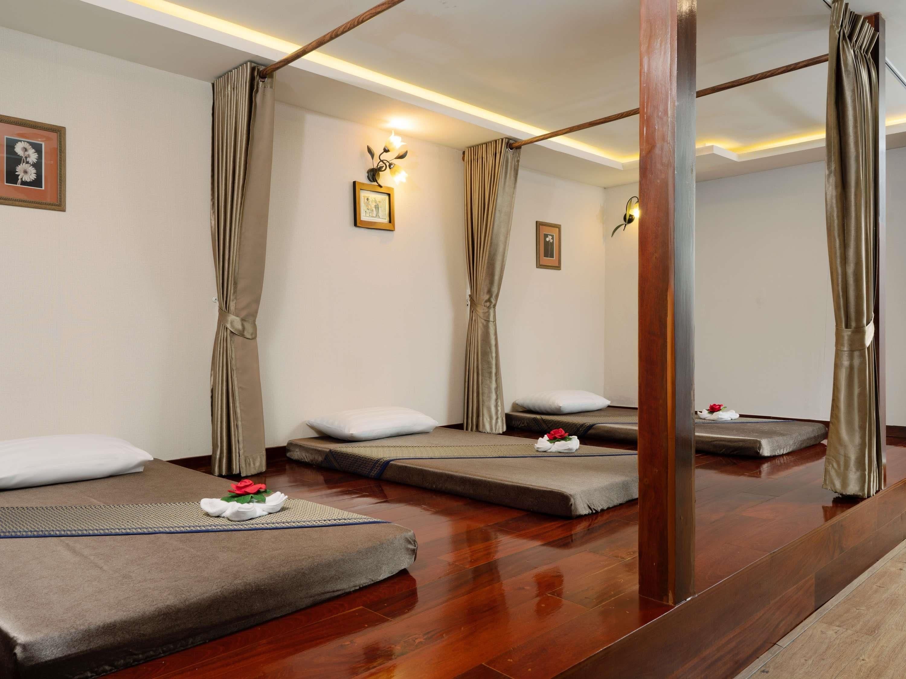 A hotel room with three single beds, each with a pillow and a towel with a flower. There are wall-mounted lights and a framed picture on the wall.