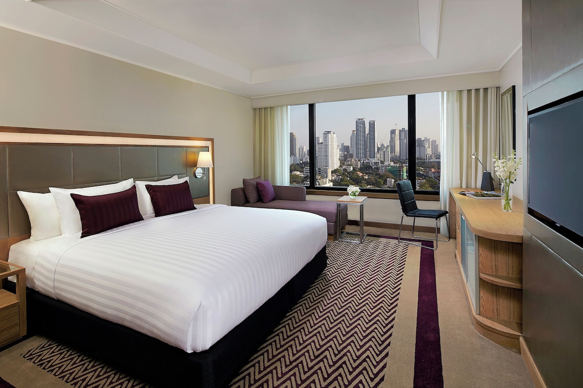A hotel room with a large bed, a desk, a chair, and a view of the city skyline.