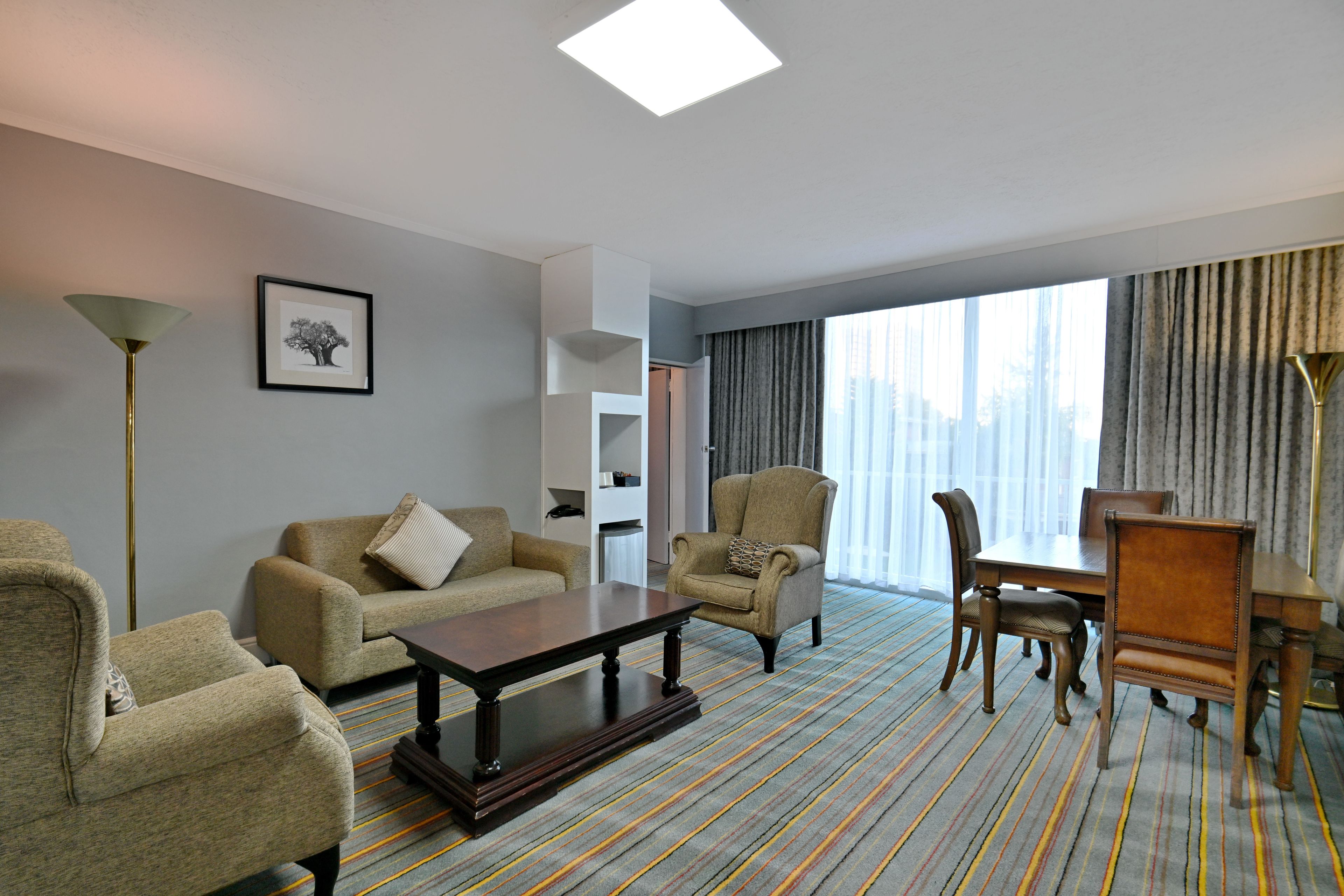 A hotel room with a sofa, armchairs, a dining table, and a large window with curtains.