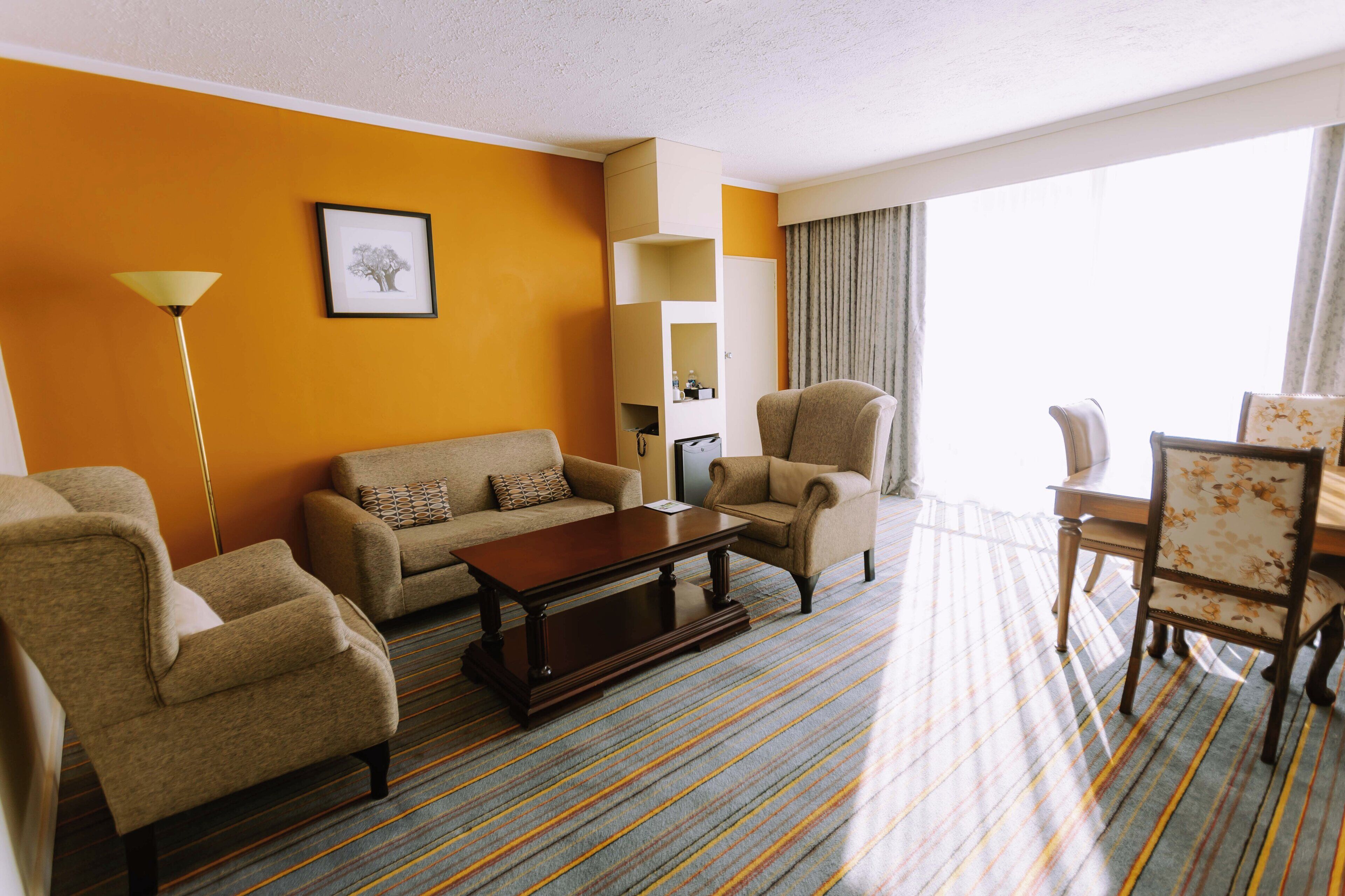 A hotel room with a sofa, armchairs, a coffee table, a dining table with chairs, a lamp, and a framed picture on the wall.