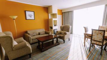A hotel room with a sofa, armchairs, a coffee table, a dining table with chairs, a lamp, and a framed picture on the wall.