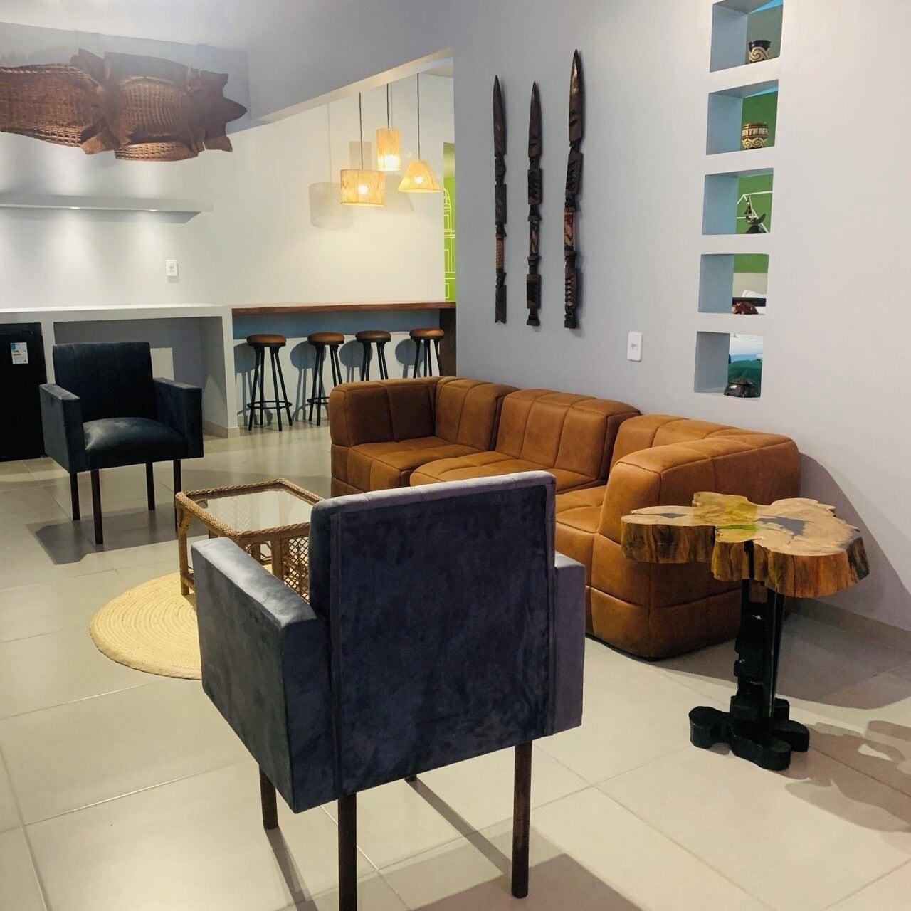 Een moderne woonkamer met een bruine leren bank, een donkerblauwe fauteuil en een ronde houten salontafel. Er zijn hangende pendellampen en een wandplank met decoratieve objecten.