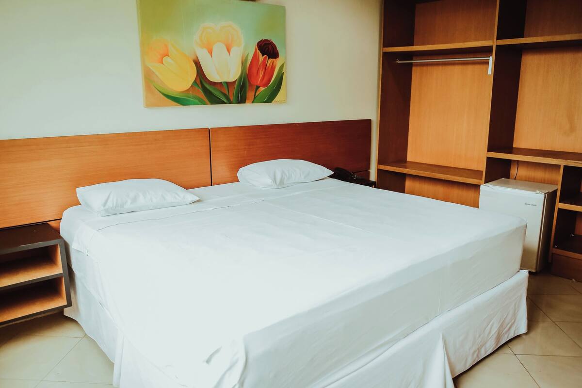 A neatly made bed with white linens, a wooden headboard, and a painting of flowers on the wall.