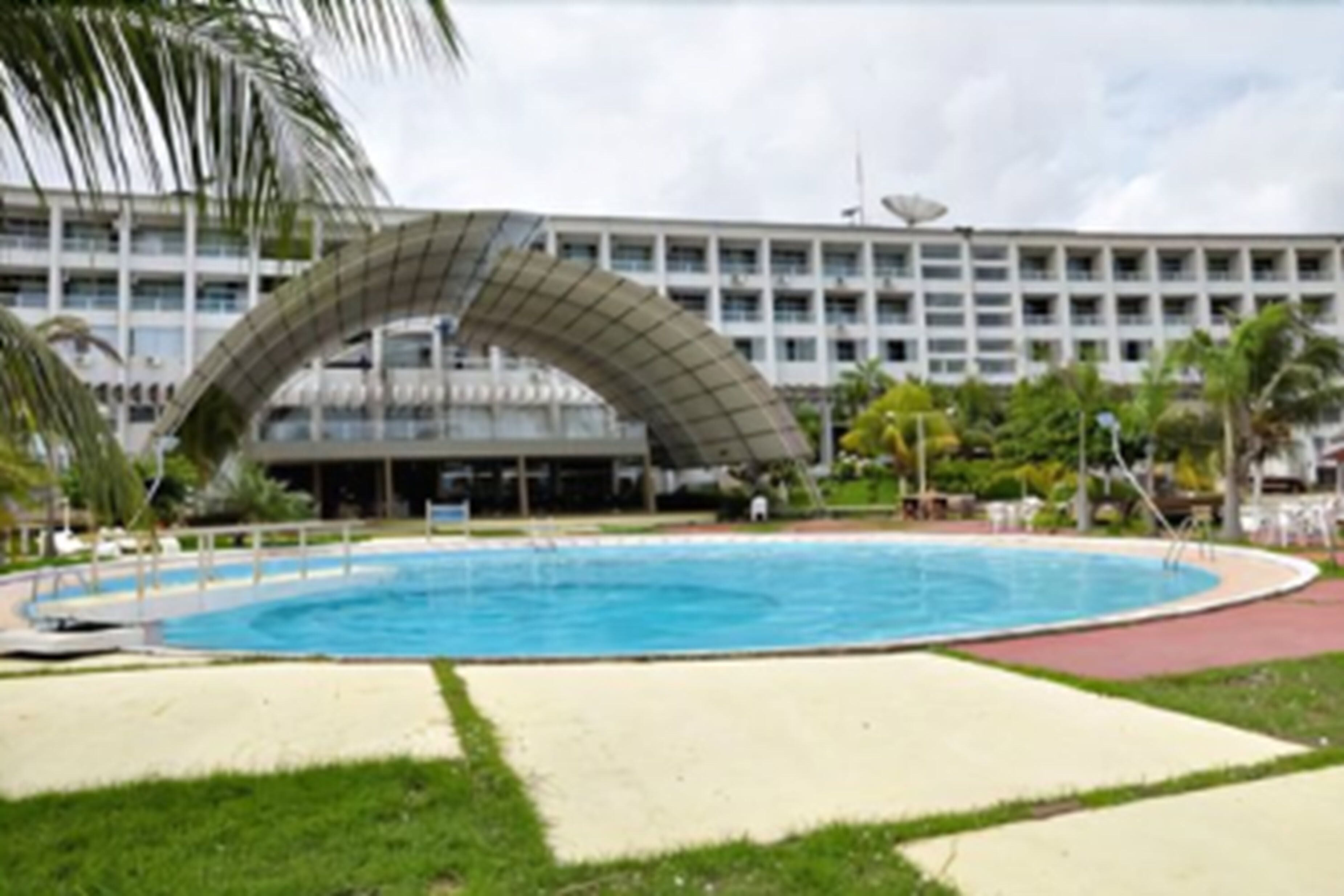 Una piscina con estructura de techo curva, rodeada de césped verde y un edificio de varios pisos al fondo.