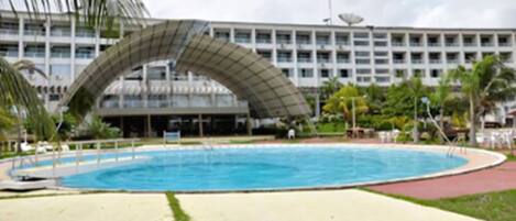 Una piscina con estructura de techo curva, rodeada de césped verde y un edificio de varios pisos al fondo.