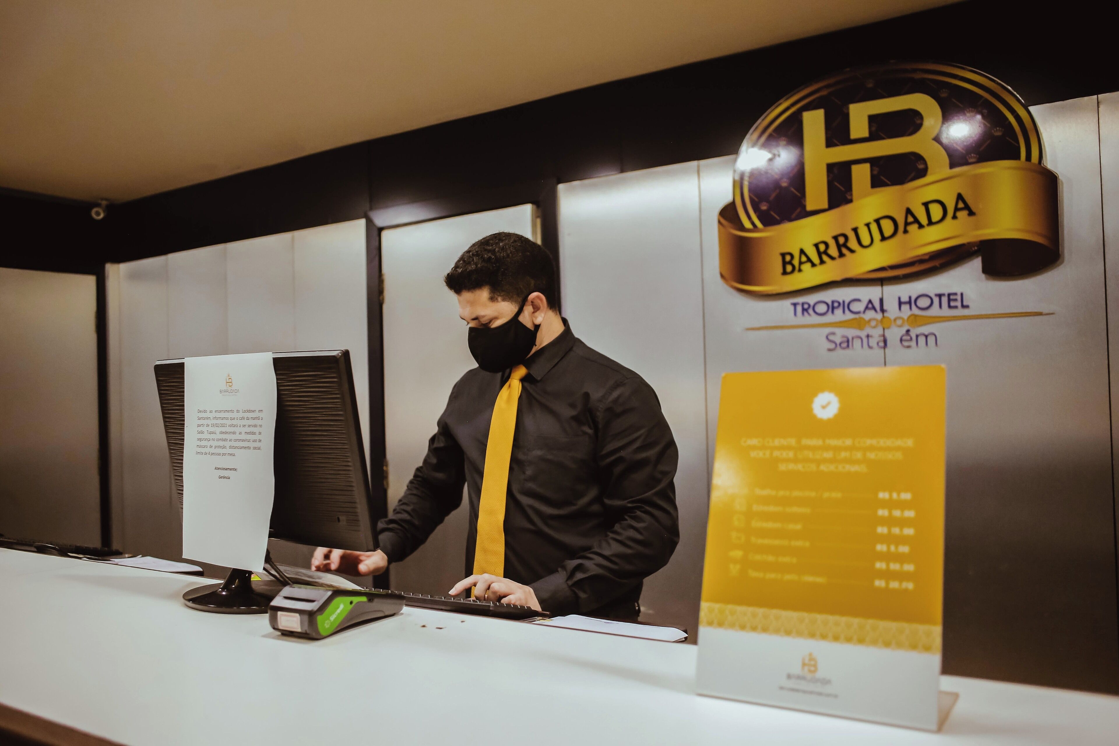 A hotel reception desk with a man in a black shirt and yellow tie. The hotel name 'BARRUDADA' and 'TROPICAL HOTEL' are visible.