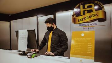 A hotel reception desk with a man in a black shirt and yellow tie. The hotel name 'BARRUDADA' and 'TROPICAL HOTEL' are visible.
