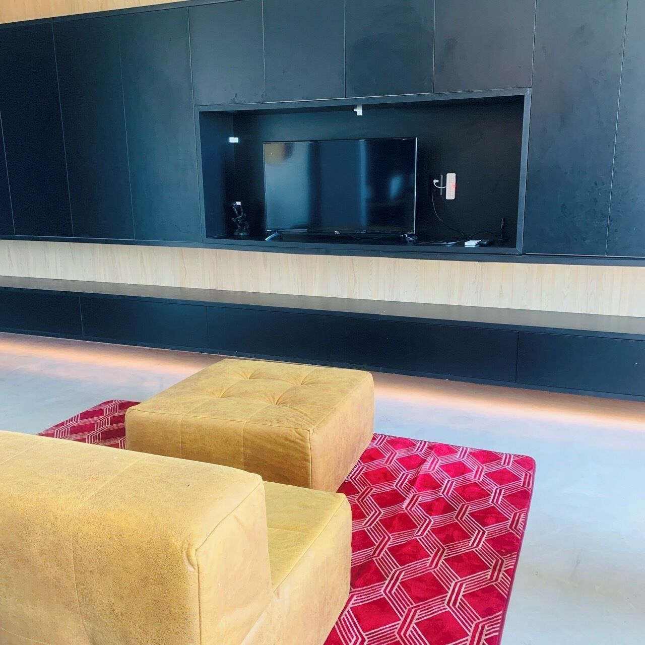 A modern living room with a flat-screen TV mounted on a dark wall, two beige ottomans, and a red patterned rug.