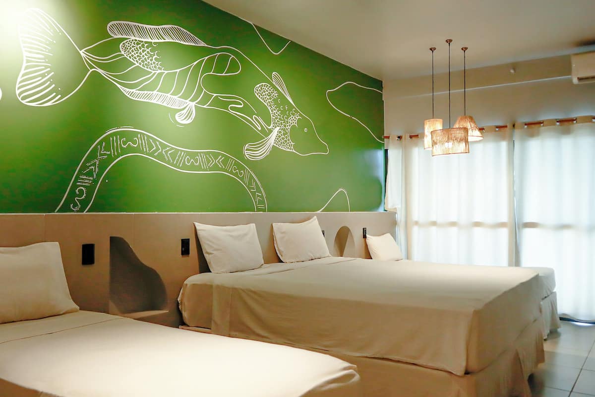 A hotel room with two beds, a green wall with white artwork, and a large window with curtains.