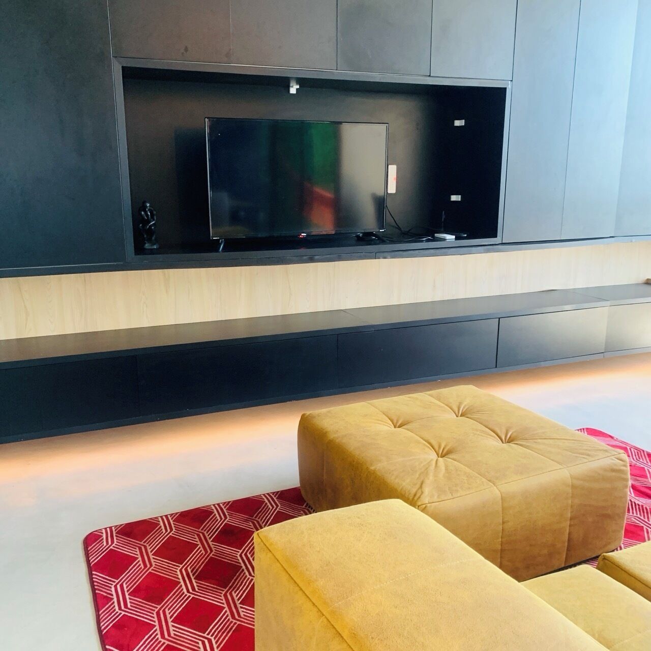 A modern living room with a flat-screen TV mounted on a dark wall, a wooden cabinet below, and two yellow ottomans on a red patterned rug.