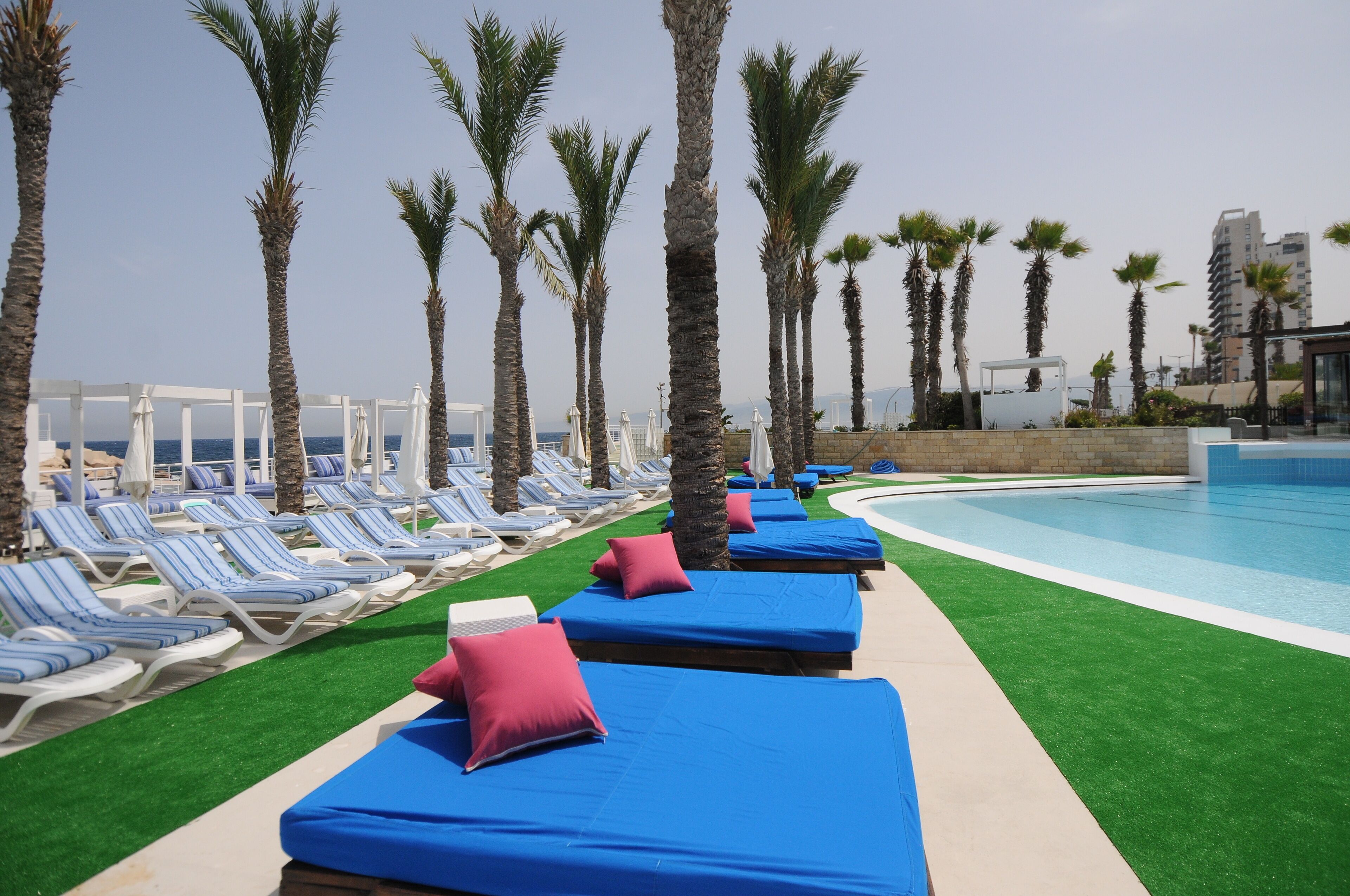 A poolside area with lounge chairs and blue mats, palm trees, and a clear sky.