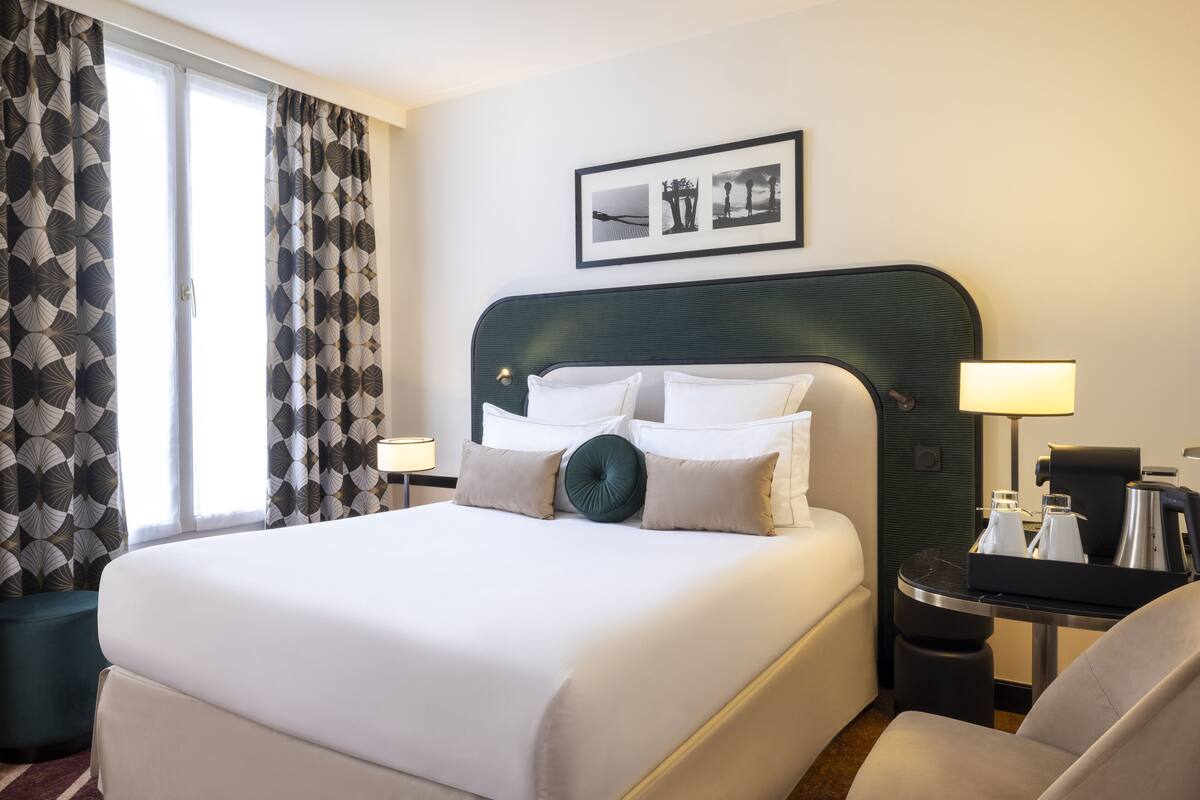 A hotel room with a large bed, a headboard, a bedside table with a lamp, a chair, and a framed picture on the wall.