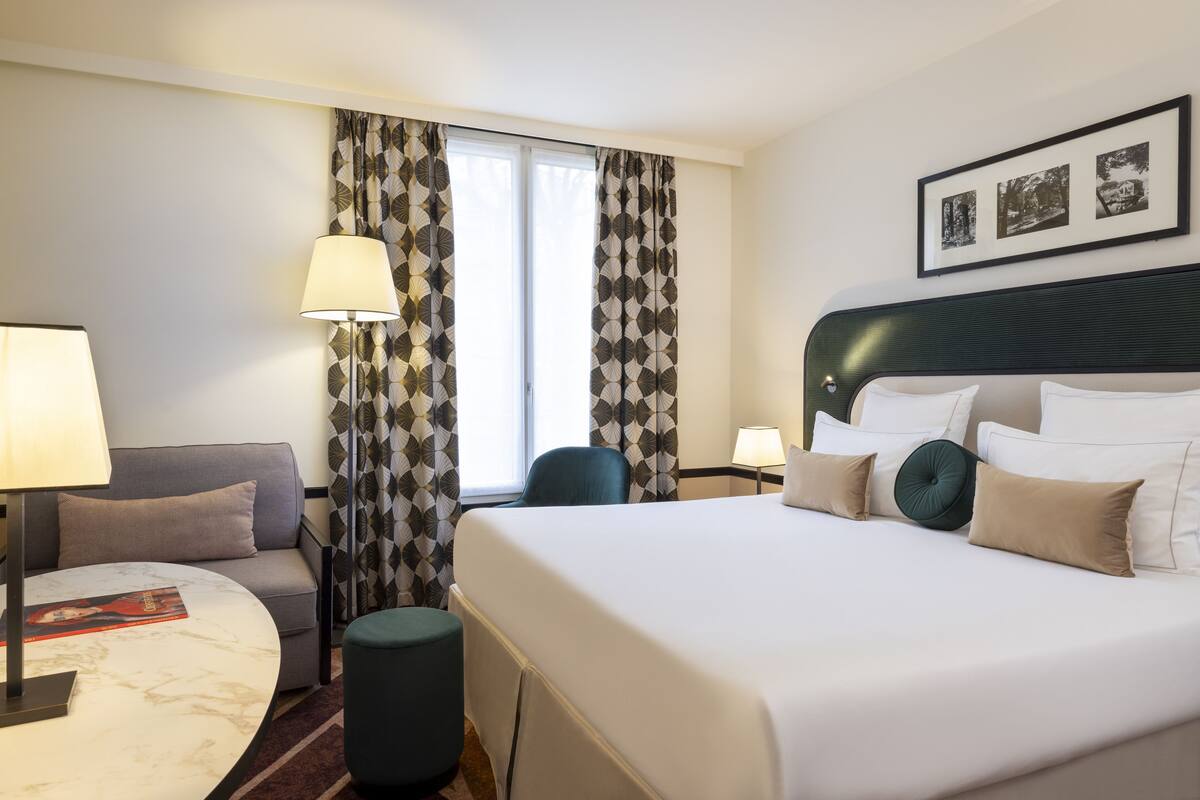 A hotel room with a large bed, a sofa, a round table, and a chair. There are two lamps, a window with curtains, and a framed picture on the wall.