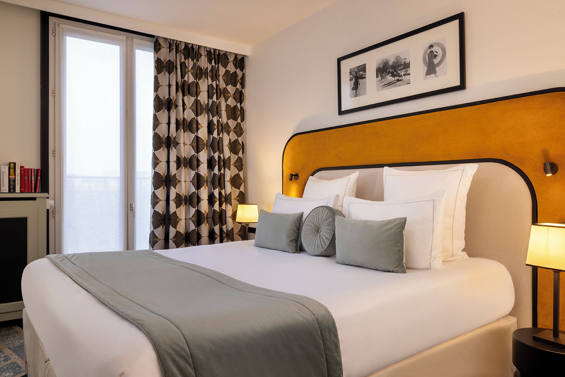 A hotel room with a bed, a headboard, a nightstand, a lamp, a window with curtains, and a framed picture on the wall.