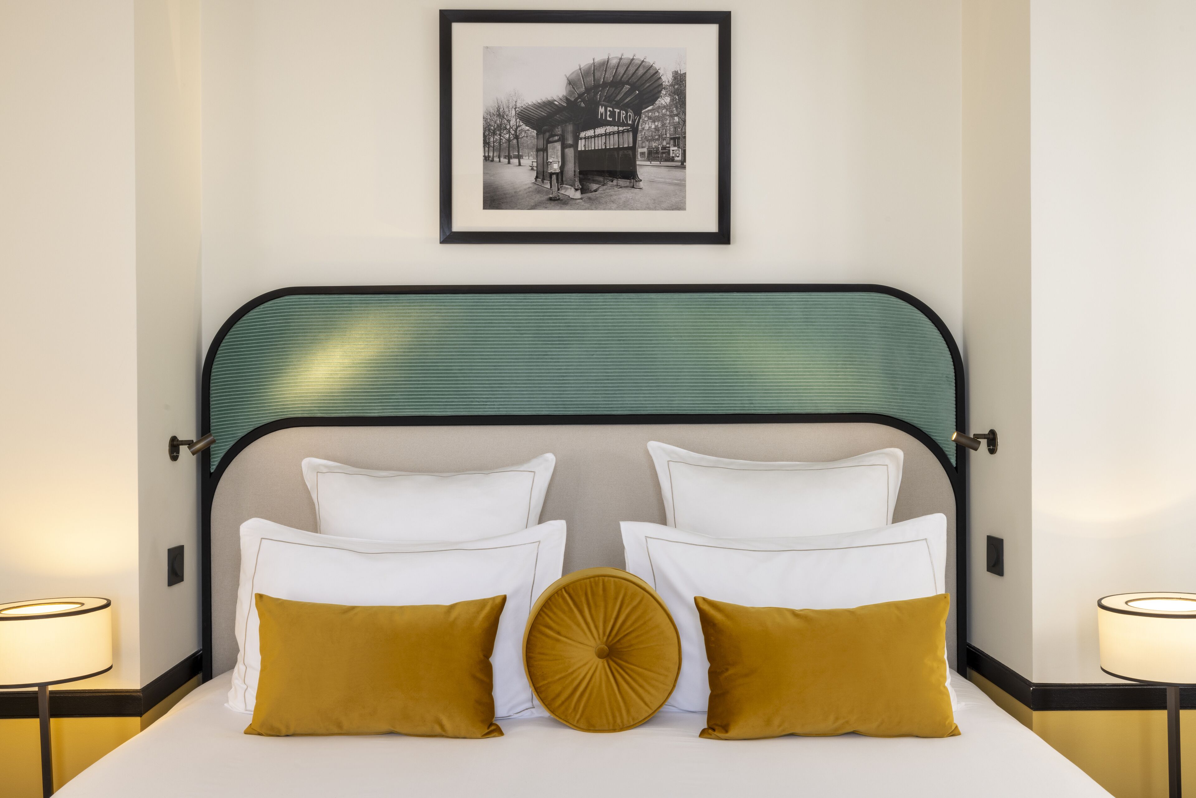 Una cama bien tendida con almohadas blancas y mostaza, un cabecero verde y un cuadro enmarcado encima.