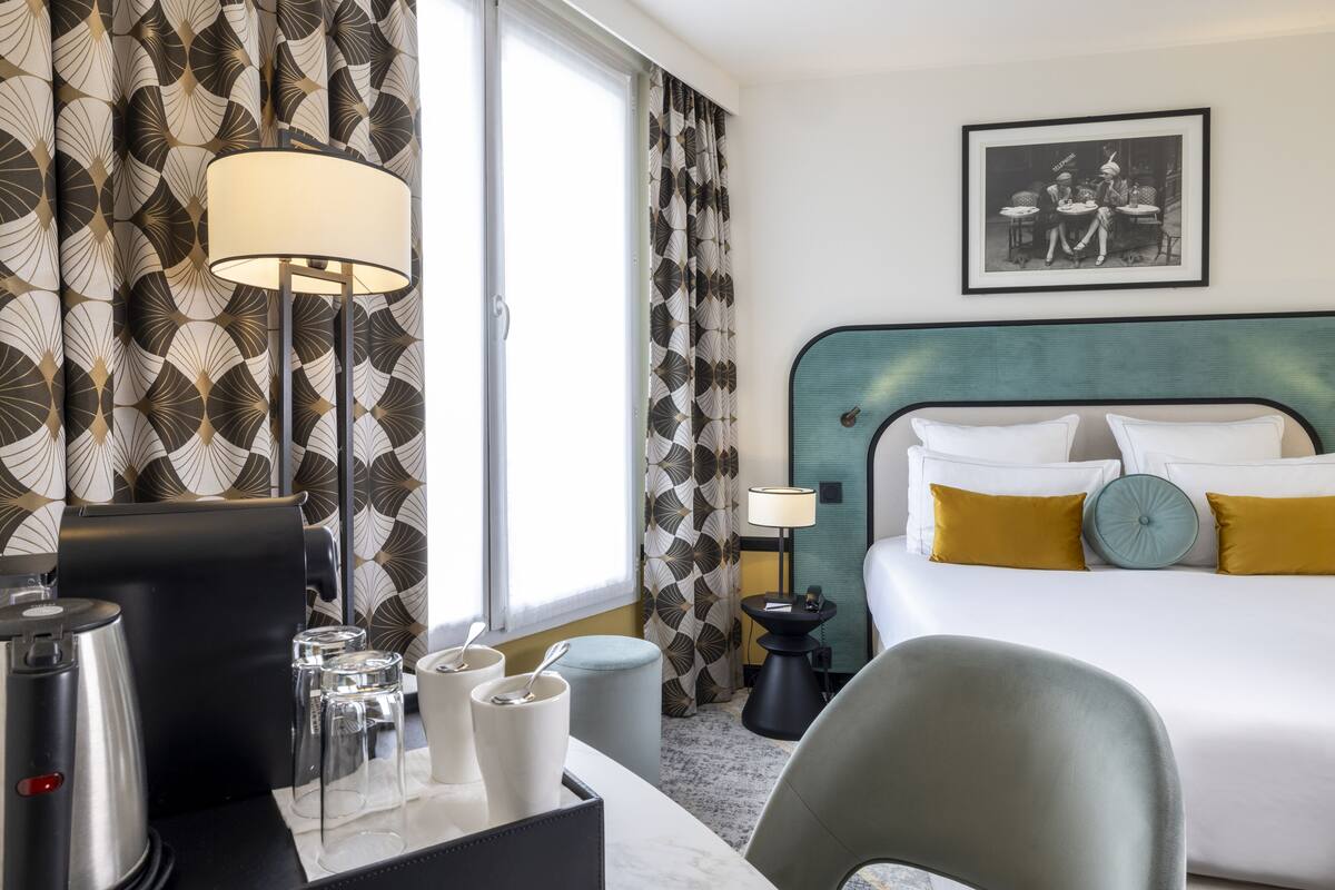 A hotel room with a bed, a desk with a coffee maker, a lamp, and a framed picture on the wall.