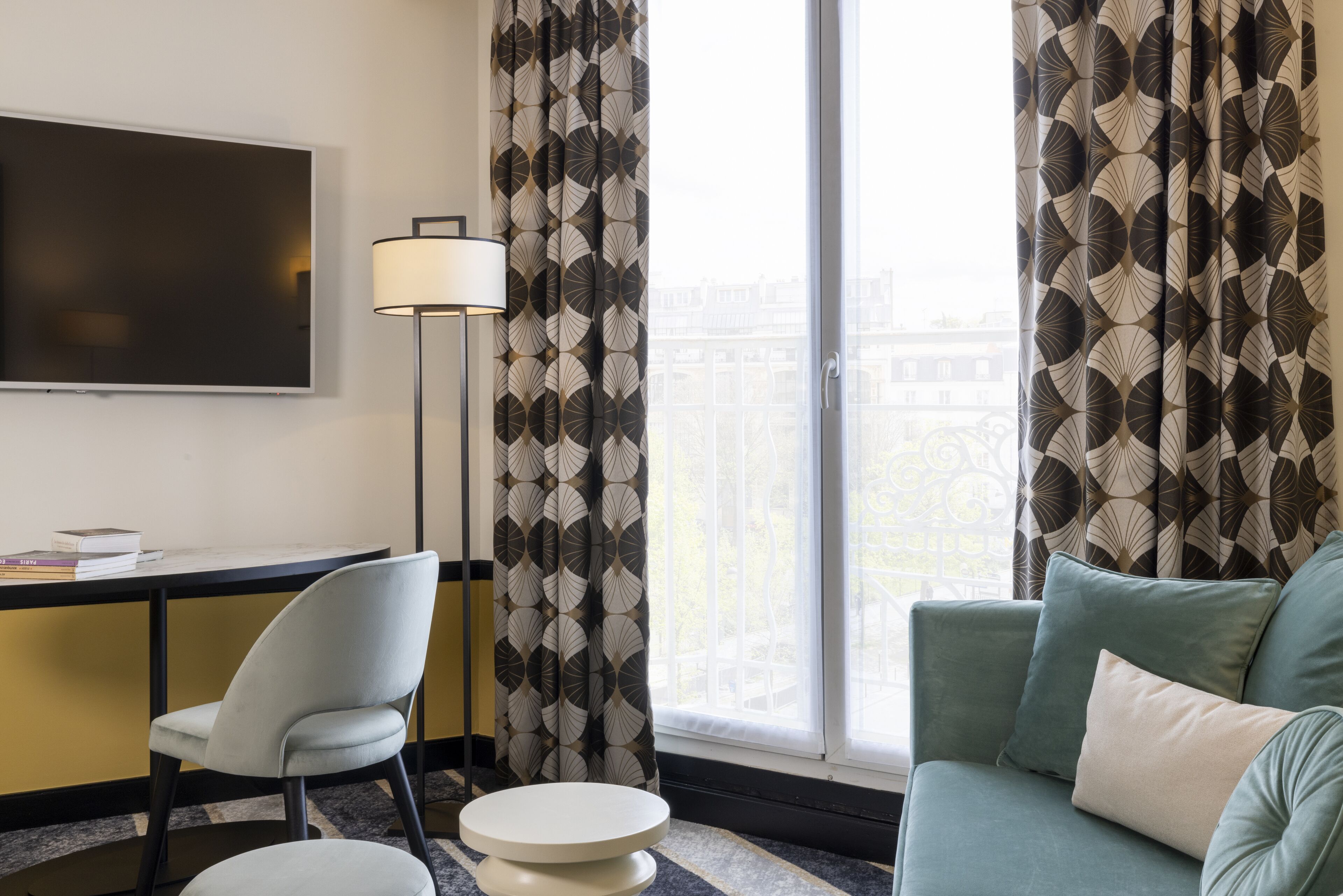 A modern hotel room with a flat-screen TV, a desk, a chair, a sofa, and patterned curtains.