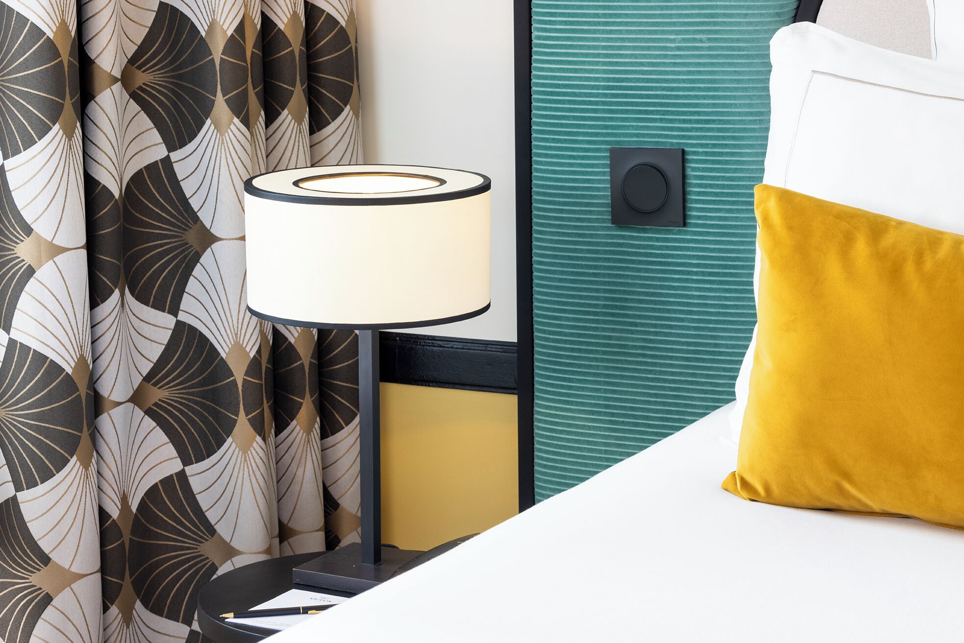 A neatly arranged hotel room with a bed, a bedside lamp, and a curtain with a geometric pattern.