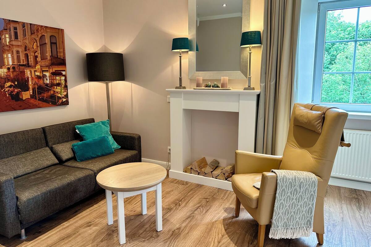 Een moderne woonkamer met een bank, een ronde houten tafel, een stoel, een open haard en een groot raam.