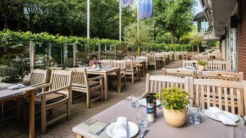 An outdoor dining area with wooden tables and chairs, surrounded by greenery and trees.